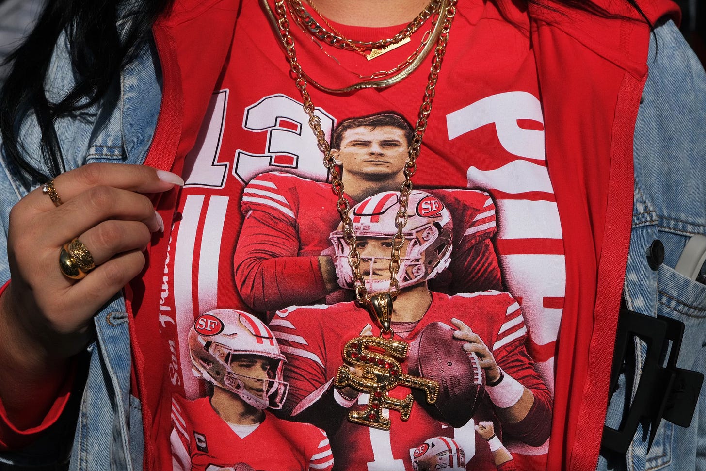 A person is wearing a red T-shirt with printed images of football players and the number 13, layered with a denim jacket and gold jewelry. A person is wearing a red T-shirt with printed images of football players and the number 13, layered with a denim jacket and gold jewelry.