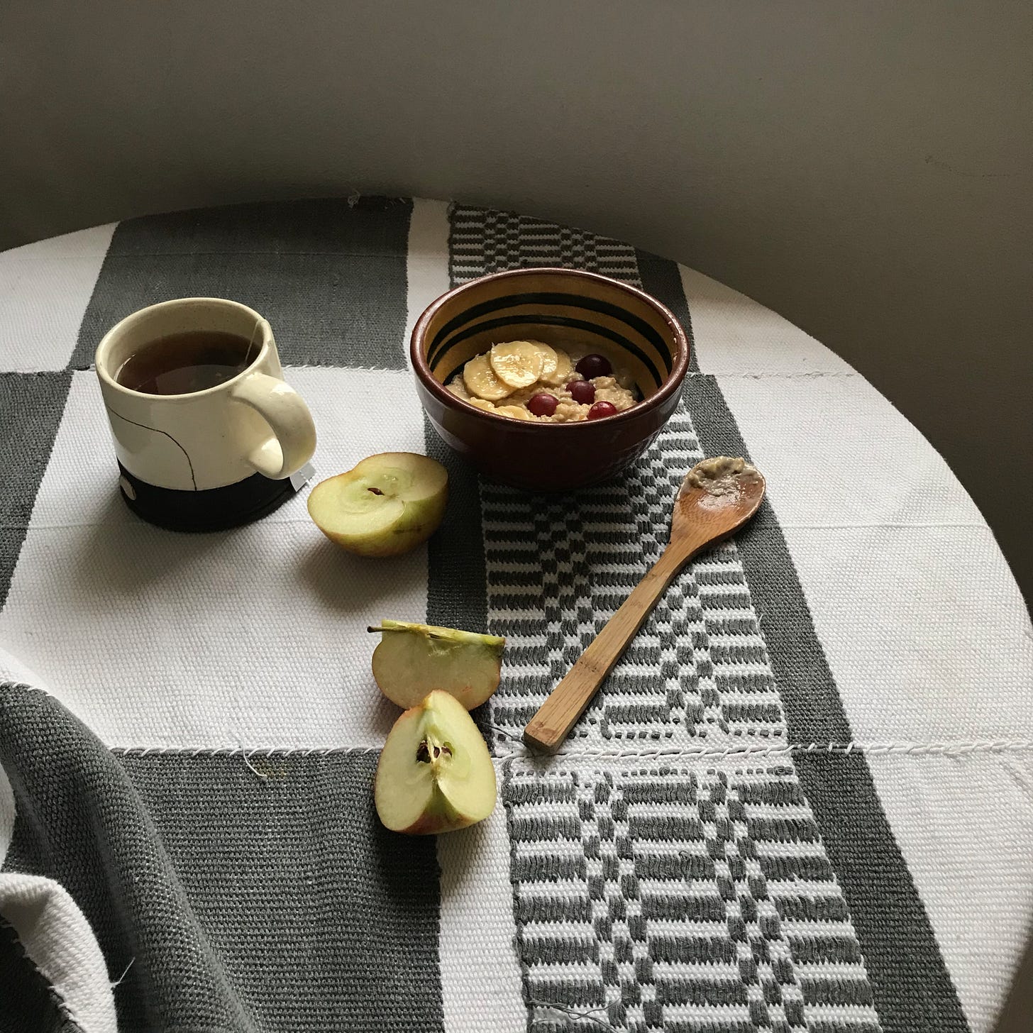 Breakfast on a round table, an apple slice into three pieces, a wooden spoon, a mug of mint tea, and a bowl of something sludgy with bananas and grapes topping it.