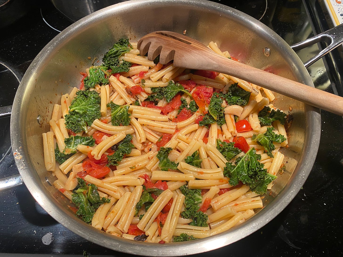 A steel pan on the stove with a wooden pasta spoon in it, stirring casarecce in a chunky, light tomato sauce with roasted garlic and kale pieces.