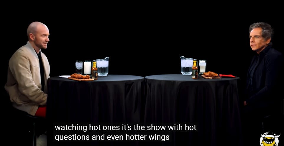 mesas redondas encostadas uma na outra, com pano de mesa preto. Cada uma comportaria duas pessoas, mas como estão grudadas colocam os dois um em cada ponto do quadro. À esquerda, Sean Evans, um homem branco e careca com uma jaqueta bege e calça vermelha, sentado em um banquinho preto. À direita, Ben Stiller, um homem branco com o cabelo se tornando grisalho e um nariz proeminente, com um olhar perdido. Ele está usando uma camisa azul e também está sentado. Na frente de cada um deles, um copo e um jarra de água, um copo de leite, uma fileira de garrafas de molho de pimenta, e asinhas de frango. Lê-se parte da legenda do vídeo: 'watching hot ones, it's the show with hot questions and even hotter wings.