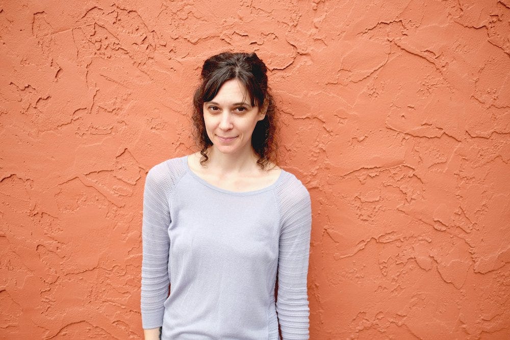 Dana is a white woman with shoulder-length dark hair, wearing a light blue top and standing against a terracotta wall. 