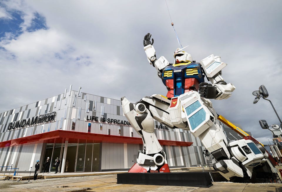 A life-sized robot statue kneels in front of a building with its right arm extended up toward the sky. Signs on the building read, "Gundam Next Future Pavilion" and "Life Is Spreading into the Universe." A life-sized robot statue kneels in front of a building with its right arm extended up toward the sky. Signs on the building read, "Gundam Next Future Pavilion" and "Life Is Spreading into the Universe."