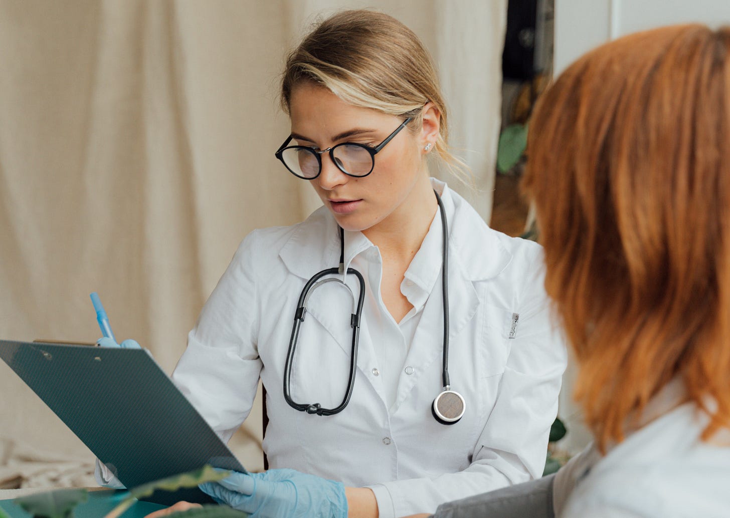 A doctor looks at a clipboard. Her patient sits in the foreground, back to the camera.
