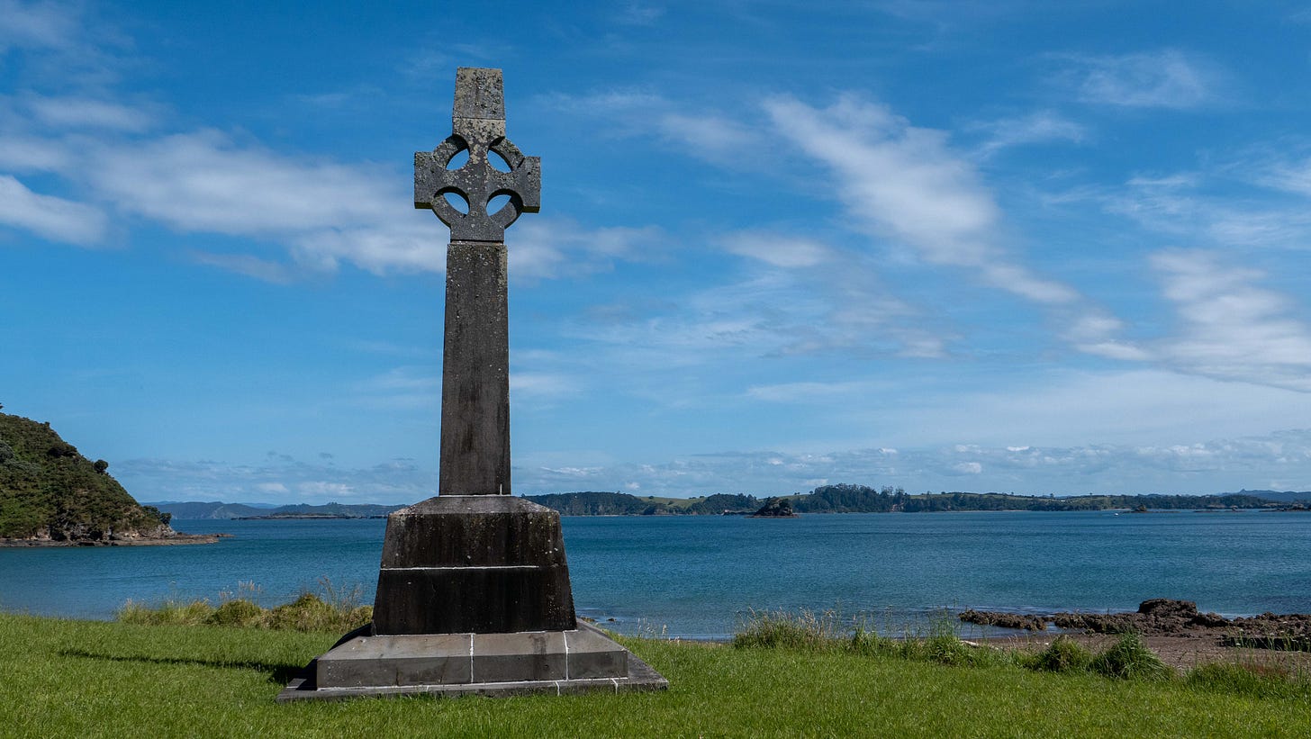 A stone cross standing in front of deep blue sea, with land behind.