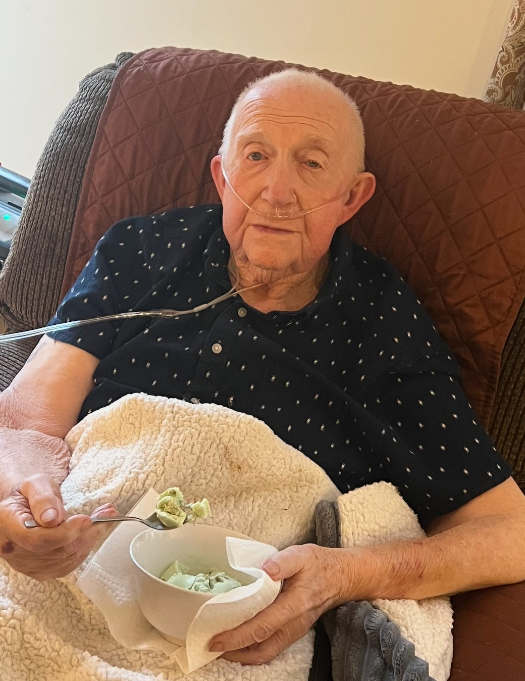 An old white man with a breathing machine sits in a chair and eats from a bowl of green pistachio ice cream