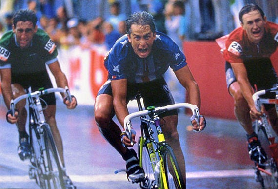 Three bicycle racers cross the finish line of the 1989 World Championships. Greg Lemond in first, wearing the stars and stripes of Team USA, his face roaring with victory. To his left, the Soviet Dmitry Konyshev in red, a young rider whose face expresses the shock of having finished second. And to Lemond's right, Irishman Sean Kelly in green, dejected at having lost a race he could have run. It is a wet day. Noe of the riders have helmets; their hair and jerseys and arms are wet. The bicycles are leaning as the riders throw their bikes towards the finish.