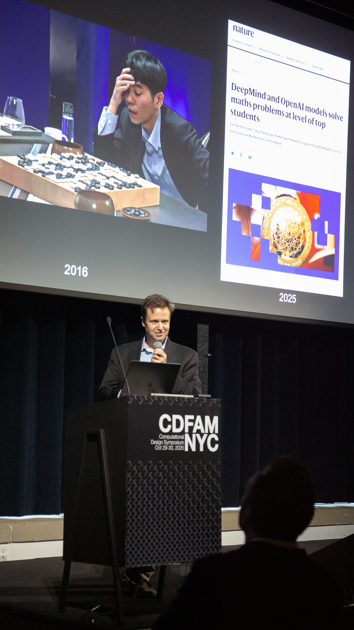 A speaker presenting at the CDFAM NYC 2025 event, showing visuals of a Go game on a screen alongside an article about AI advancements in solving math problems.