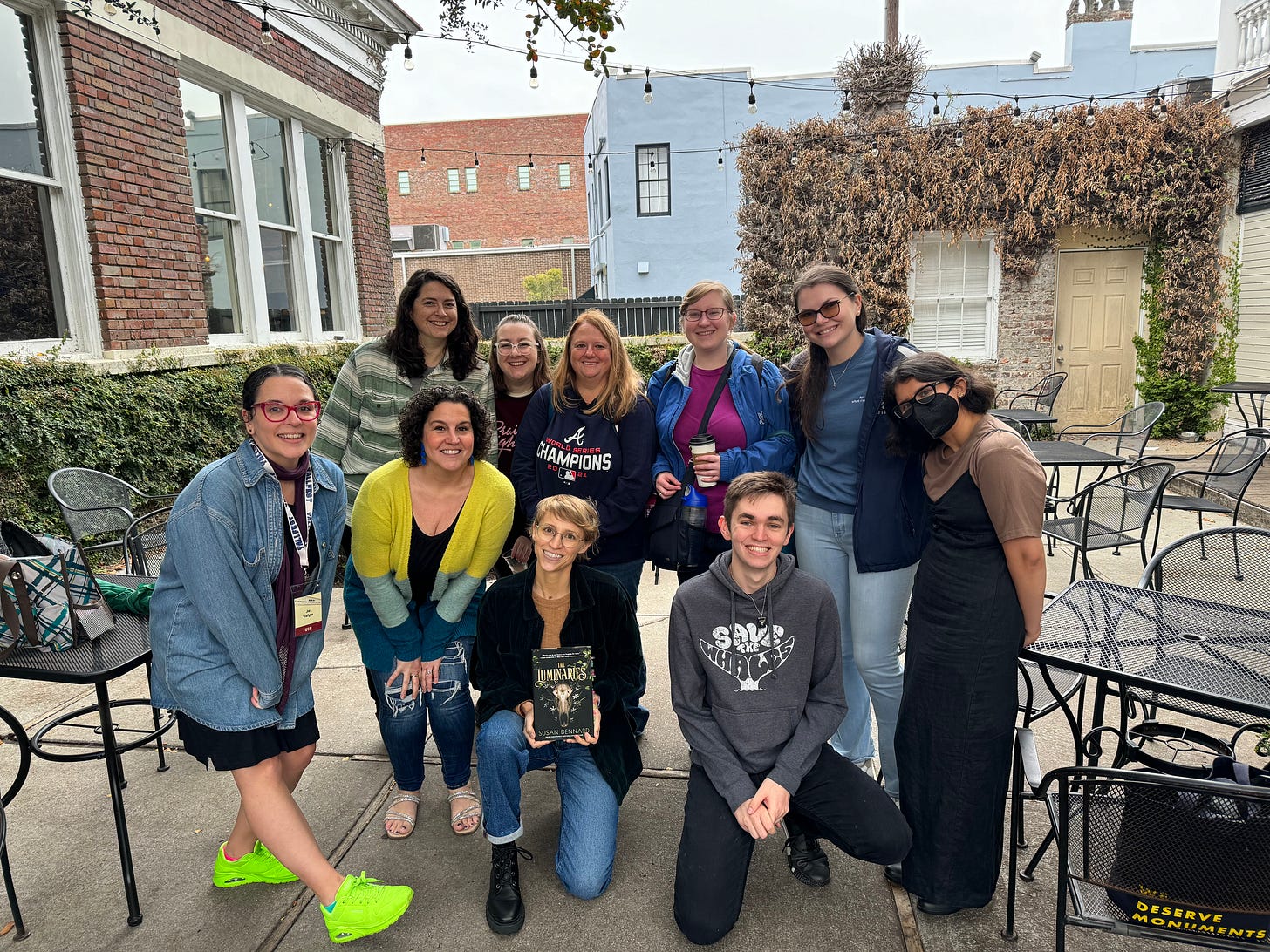 A photo of Sooz surrounded by readers at YALLFest. Everyone is smiling, and she's holding a copy of The Luminaries A photo of Sooz surrounded by readers at YALLFest. Everyone is smiling, and she's holding a copy of The Luminaries
