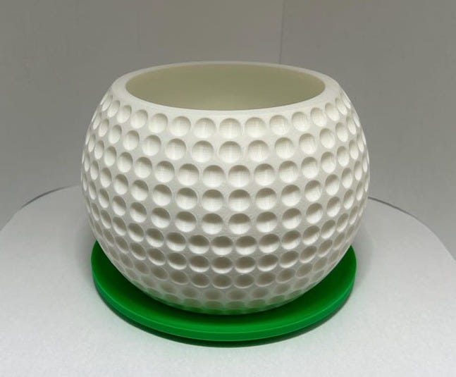 May include: A white golf ball-textured planter with a green saucer. The planter is round and has a dimpled surface, resembling a golf ball. It's made of a smooth, matte white material. The green saucer provides a contrasting base. This decorative planter is perfect for adding a touch of sporty style to any indoor or outdoor space.  It's ideal for small plants, succulents, or air plants.