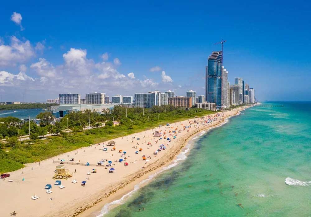 A wide aerial view of Haulover Beach in Miami, Florida, showing a long stretch of sandy shoreline with beachgoers under umbrellas, turquoise ocean waves on the right, and high-rise buildings lining the coast in the background. A wide aerial view of Haulover Beach in Miami, Florida, showing a long stretch of sandy shoreline with beachgoers under umbrellas, turquoise ocean waves on the right, and high-rise buildings lining the coast in the background.