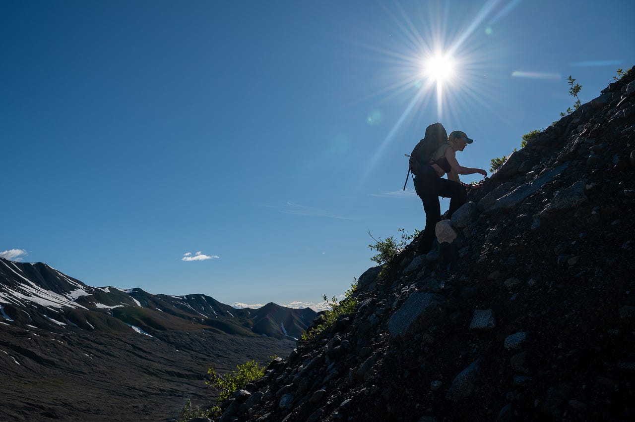 Backlit by a bright sunburst at the top of the frame, a hiker with a pack is silhouetted climbing a steep slope of dark scree, one hand reaching to the rocks. Beyond and below, snow-streaked ridges fall away into a wide valley under a deep blue sky.