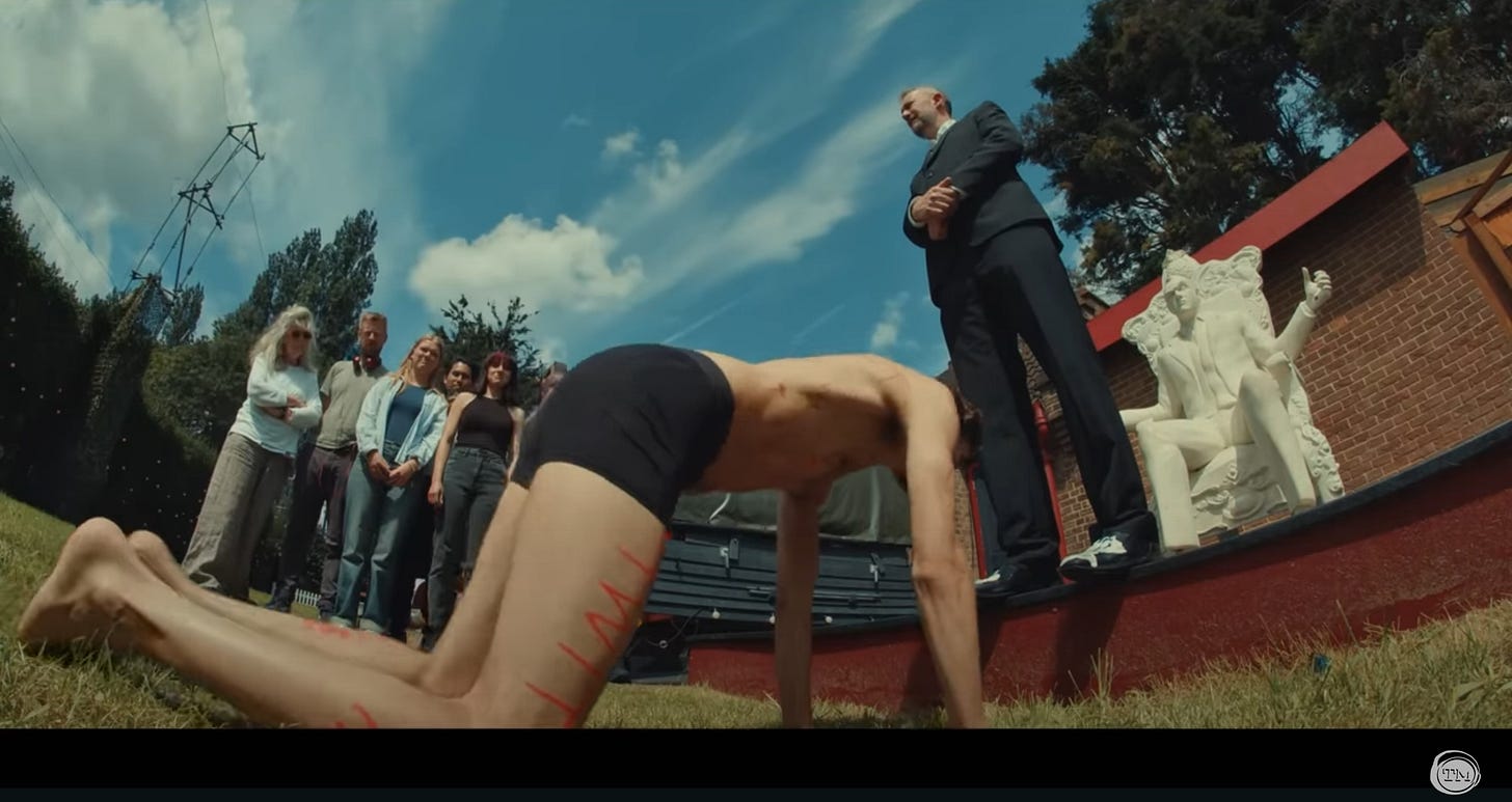 Mathew Baynton, wearing only boxer briefs, gets on his hands and knees to kick yogurt off Little Alex Horne’s shoes, as a small group of people watch.