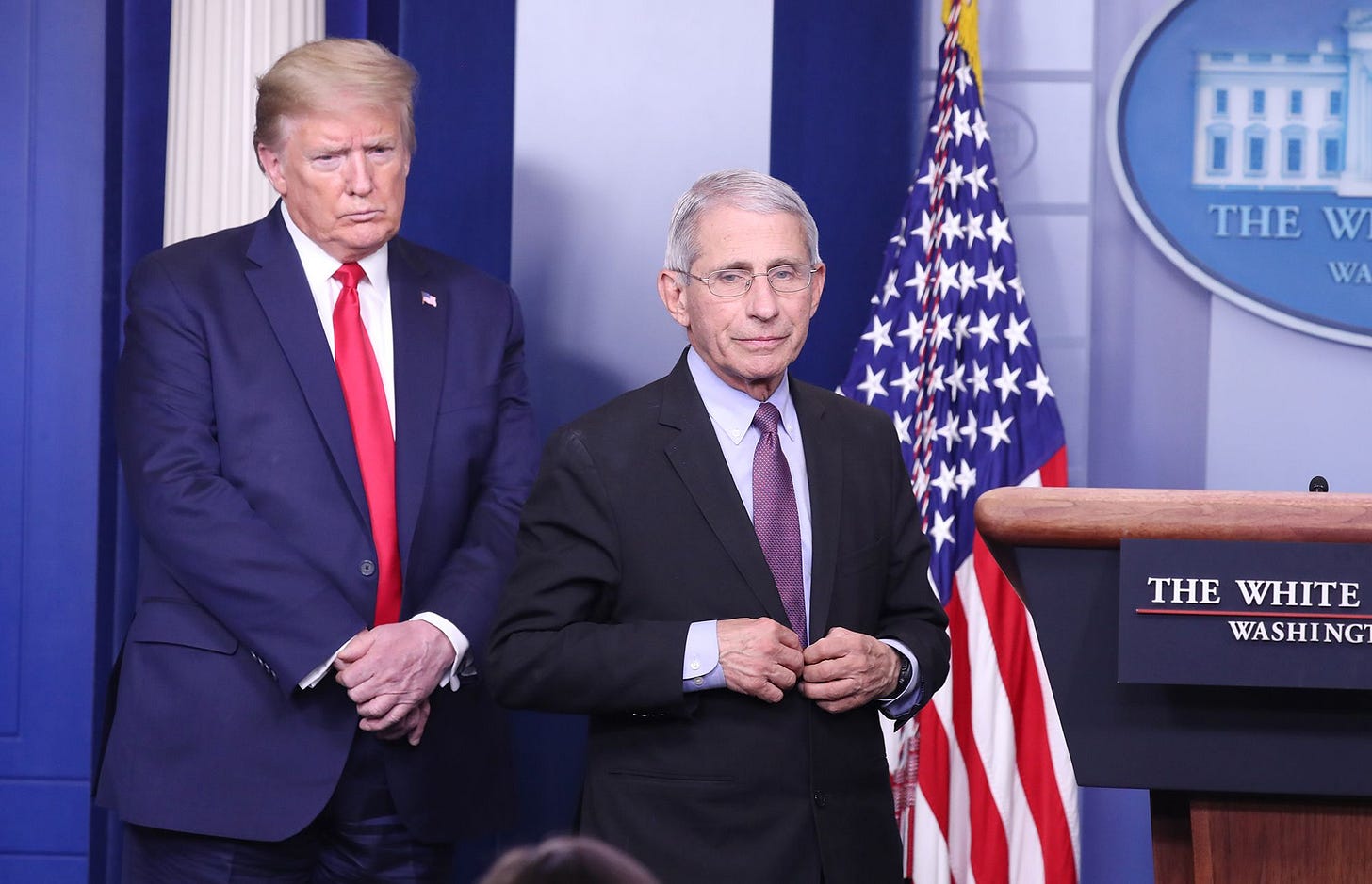 Dr. Anthony Fauci stands onstage at the White House briefing room with Trump behind him.