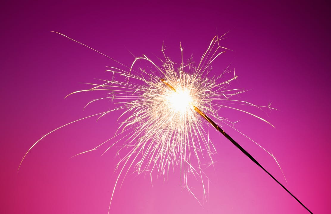 A single lit sparkler flares white-hot against a deep pink background, flinging golden sparks in every direction. A single lit sparkler flares white-hot against a deep pink background, flinging golden sparks in every direction.