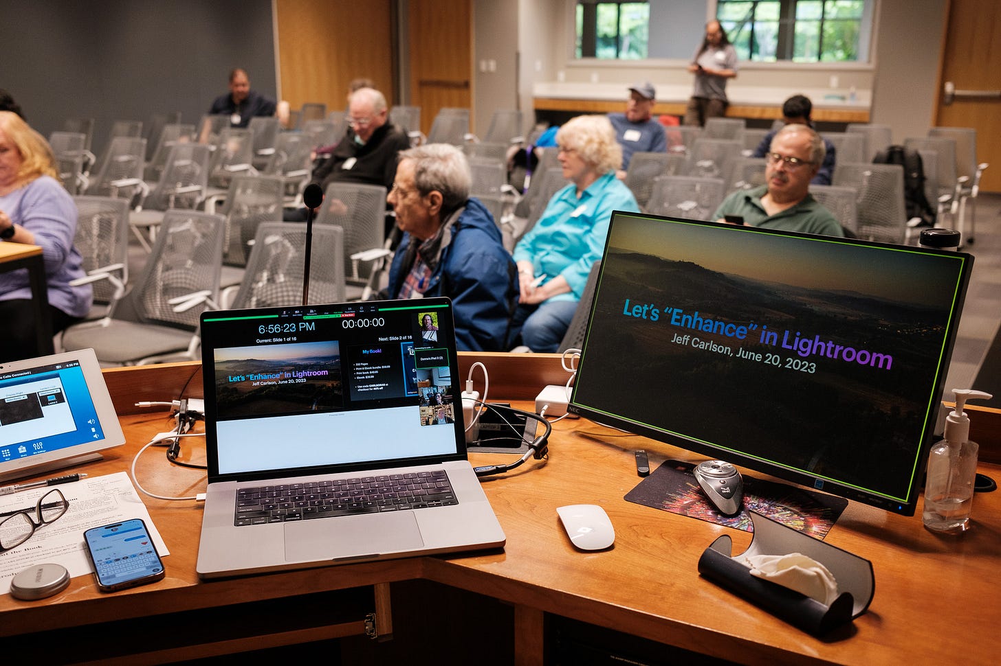 A laptop computer connected to an external display at a lectern in a conference room with a few early attendees seated in chairs.