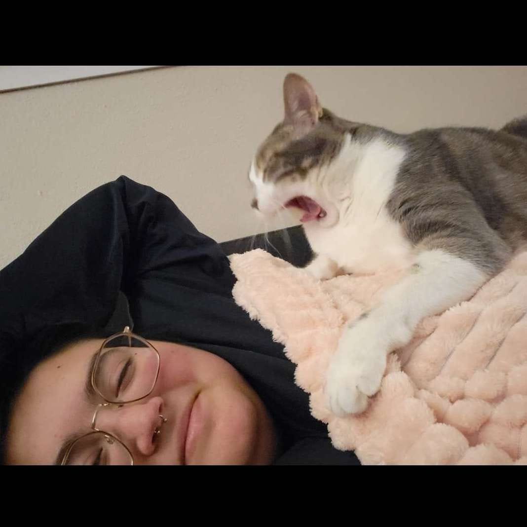 Me laying on a couch with Lorca the cat laying on top of me. She's yawning and i'm laughing