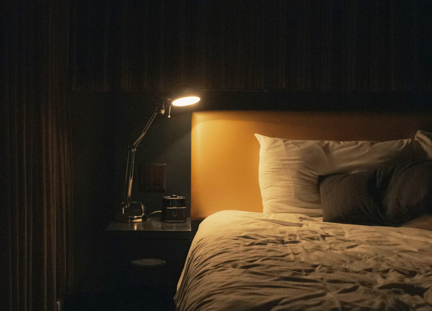 Warm light falls across an unmade bed in a quiet, dimly lit room, evoking solitude and stillness.