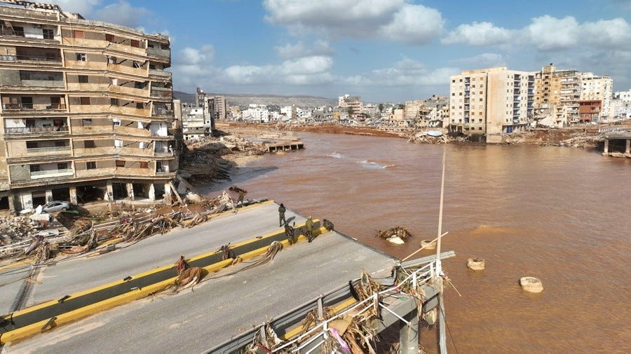 Several people stand on what remains of a road bridge that has been washed away by a flood, beside many damaged buildings. Several people stand on what remains of a road bridge that has been washed away by a flood, beside many damaged buildings.
