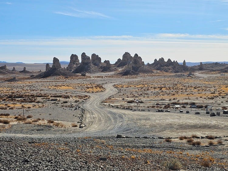 Winding dirt road through a barren desert leading toward tall, cone‑shaped rock pinnacles under a clear blue sky.