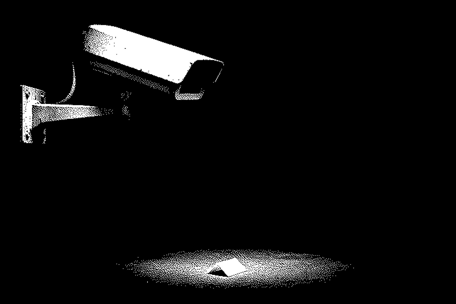 An old wall-mounted security camera on a metal bracket, seen side-on against pure black, its lens aimed off to the right. One hard light from above. Directly beneath the camera, on the bare floor in its blind spot, lies a single small folded paper no