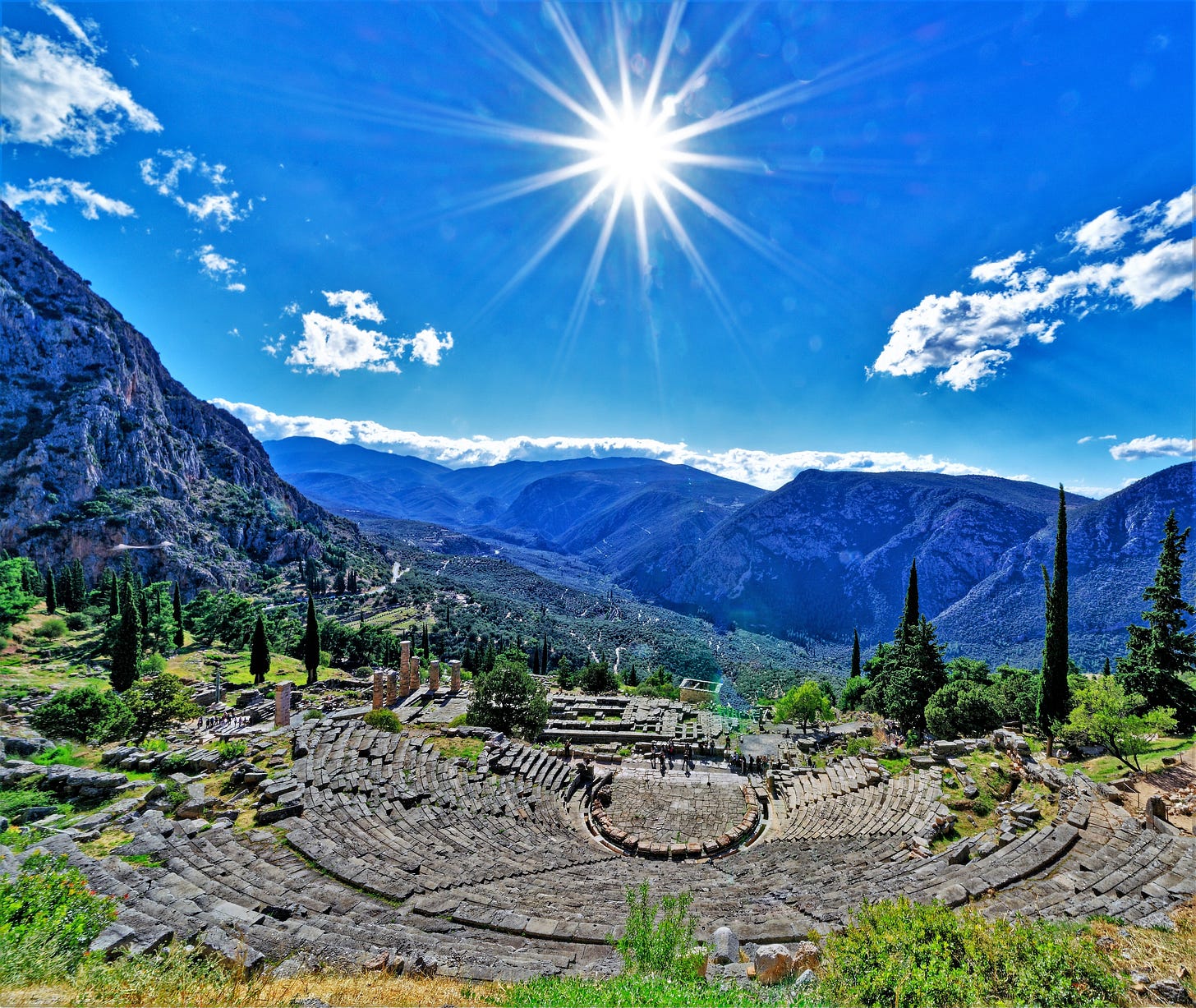 Ruins of the ancient theater at Delphi on a mountainside, with stone seating curving around the stage and Mount Parnassus rising in the background beneath a bright sun and blue sky.