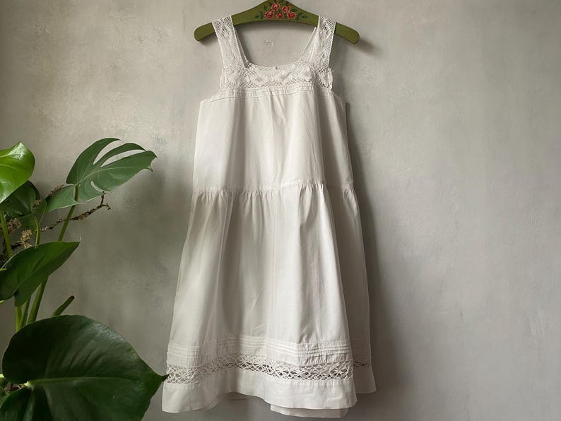 May include: A white sleeveless dress with lace trim, hanging on a green wooden hanger. The dress features a square neckline and a tiered skirt. The lace detailing is visible on the straps, neckline, and hemline. The dress is made of a lightweight fabric.