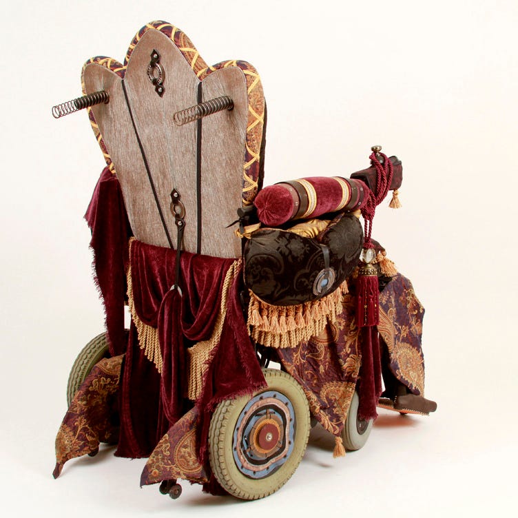 SIde back view of an electric wheelchair upholstered in silver and gold with purple and gold drapery, tassels, and fringe.