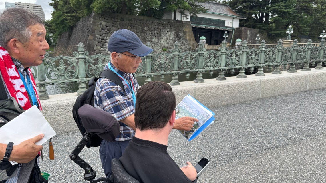Two volunteer guides show a wheelchair user pages from a guidebook beside the Imperial Palace bridge, with the moat wall and palace gate behind them. Two volunteer guides show a wheelchair user pages from a guidebook beside the Imperial Palace bridge, with the moat wall and palace gate behind them.