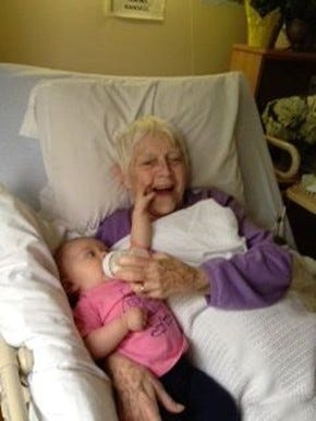Photograph of a smiling elderly woman in bed holding a baby with a bottle
