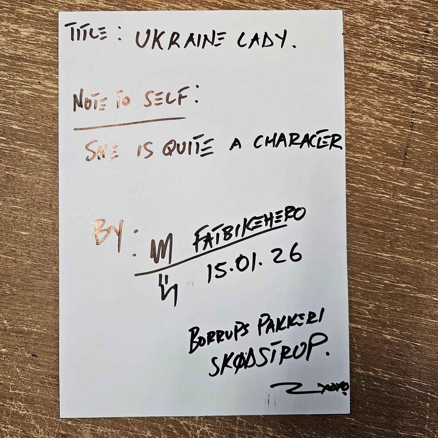 Alt Text: The back of a white card featuring handwritten black ink text. It reads: "TITLE: UKRAINE LADY. NOTE TO SELF: SHE IS QUITE A CHARACTER. BY: M FATBIKEHERO. 15.01.26. BORRUPS PAKKERI SKØDSTRUP."