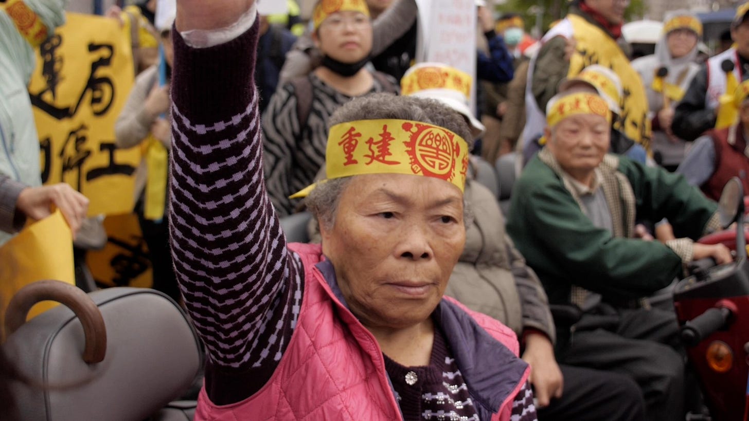 An elderly woman seated in a mobility scooter and wearing a yellow headband with red lettering raises her fist above her head in protest at the threats against Losheng Sanitorium