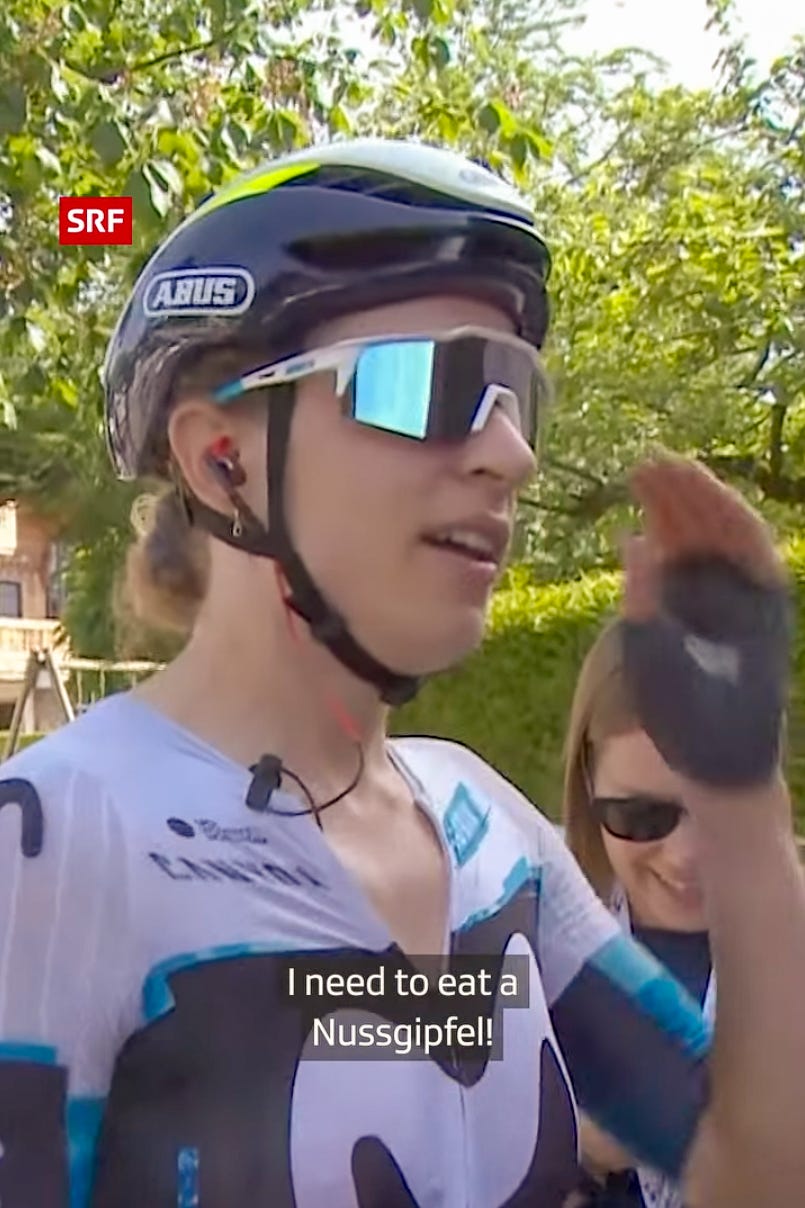 Screenshot eines SRF-Fernsehbeitrags mit der Schweizer Spitzen-Radsportlerin Marlen Reusser nach ihrem Gesamtsieg an der Tour de Suisse Women 2026. Sie trägt einen schwarzen Fahrradhelm, eine grosse verspiegelte Sportbrille und das Trikot ihres Teams. Während eines Interviews im Freien wünscht sie sich ales erstes einen Nussgipfel. Im Bild eingeblendet ist ihr Zitat: «I need to eat a Nussgipfel!».