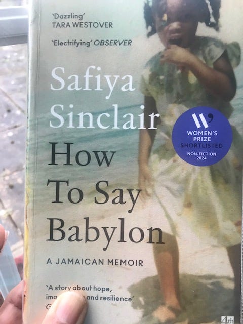 Photo of book, showing the cover of 'How To Say Babylon' by Safiya Sinclair. Image is a photo of a black Jamaican girl of about five years of age on the beach with the sea in the background. She has multiple pig tails, a light-coloured dress and brown sandals.
