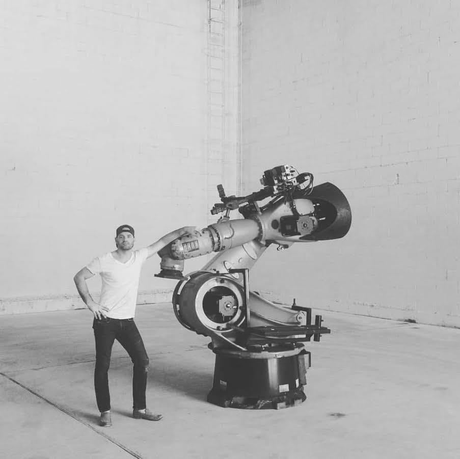Black and white photo of a man standing next to a large industrial robotic arm in an empty warehouse.