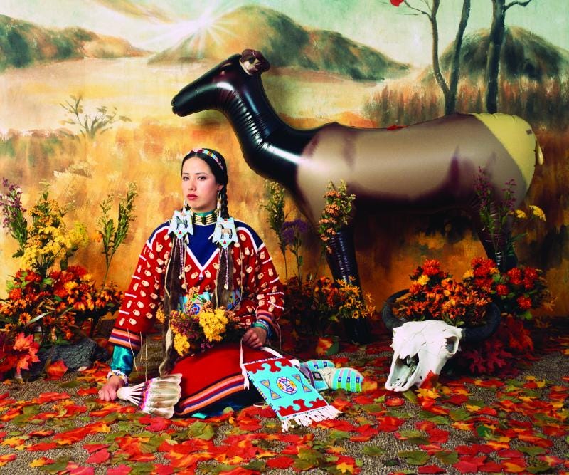 Woman in Native outfit sitting in front of a plains background with a cow skull next to her and an inflatable elk behind her. Woman in Native outfit sitting in front of a plains background with a cow skull next to her and an inflatable elk behind her.