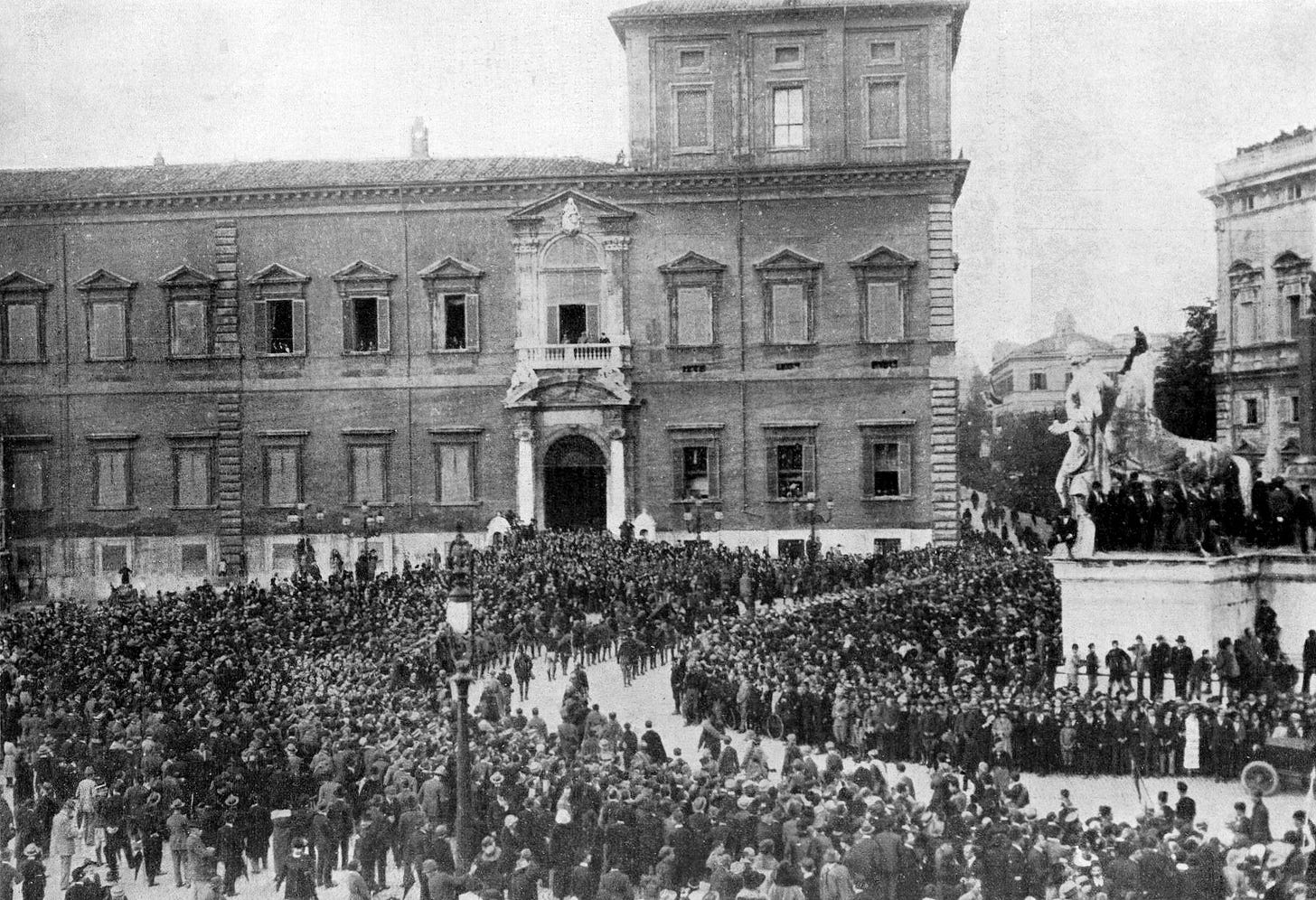 A dense crowd of men fills a Roman square in front of the Quirinal Palace, a large stone building, as columns of Fascist squads parade past during the March on Rome.