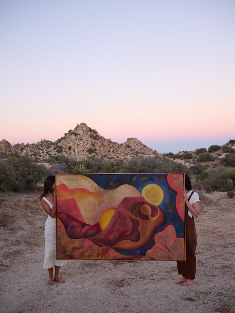 Two people hold Alex Maceda’s large abstract painting the desert will hold you in a desert landscape at dusk, with soft pink sky and rocky hills behind them. Two people hold Alex Maceda’s large abstract painting the desert will hold you in a desert landscape at dusk, with soft pink sky and rocky hills behind them.