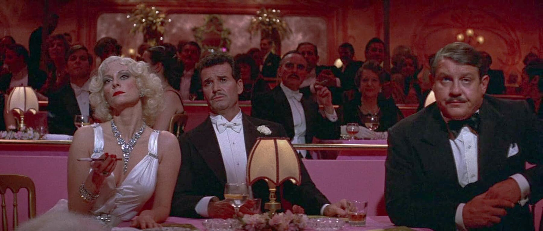 A bored Lesley Ann Warren in a white gown sits beside a skeptical James Garner, with Alex Karras to their right—Parisian nightclub patrons frozen mid-reaction.