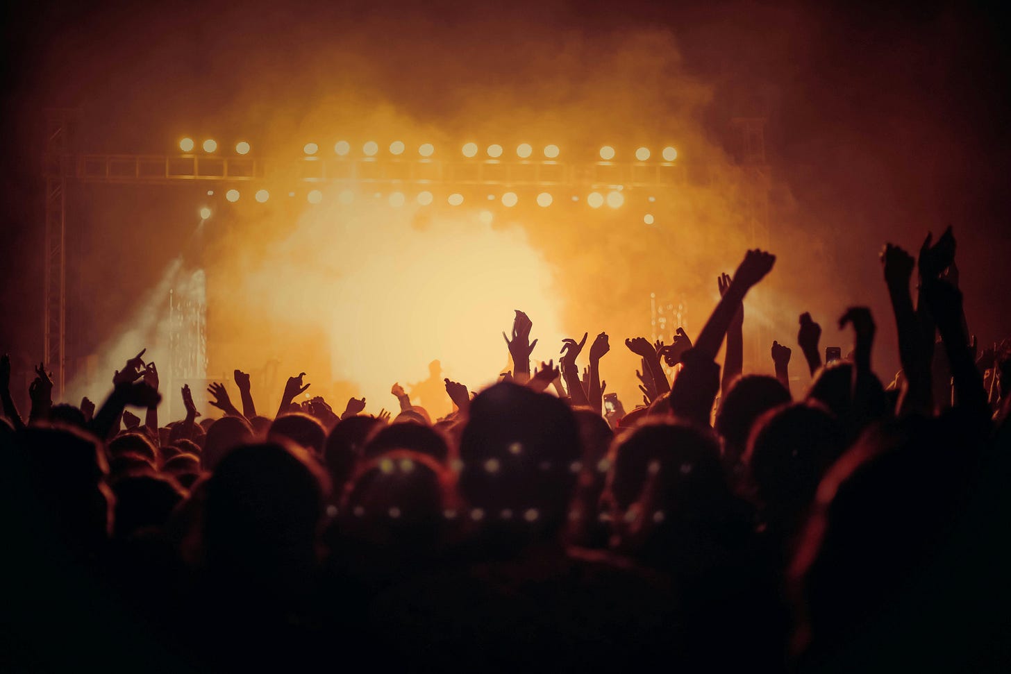 A crowd of people at an outdoor music festival at night. The people are silhouetted by yellow-orange stage lights