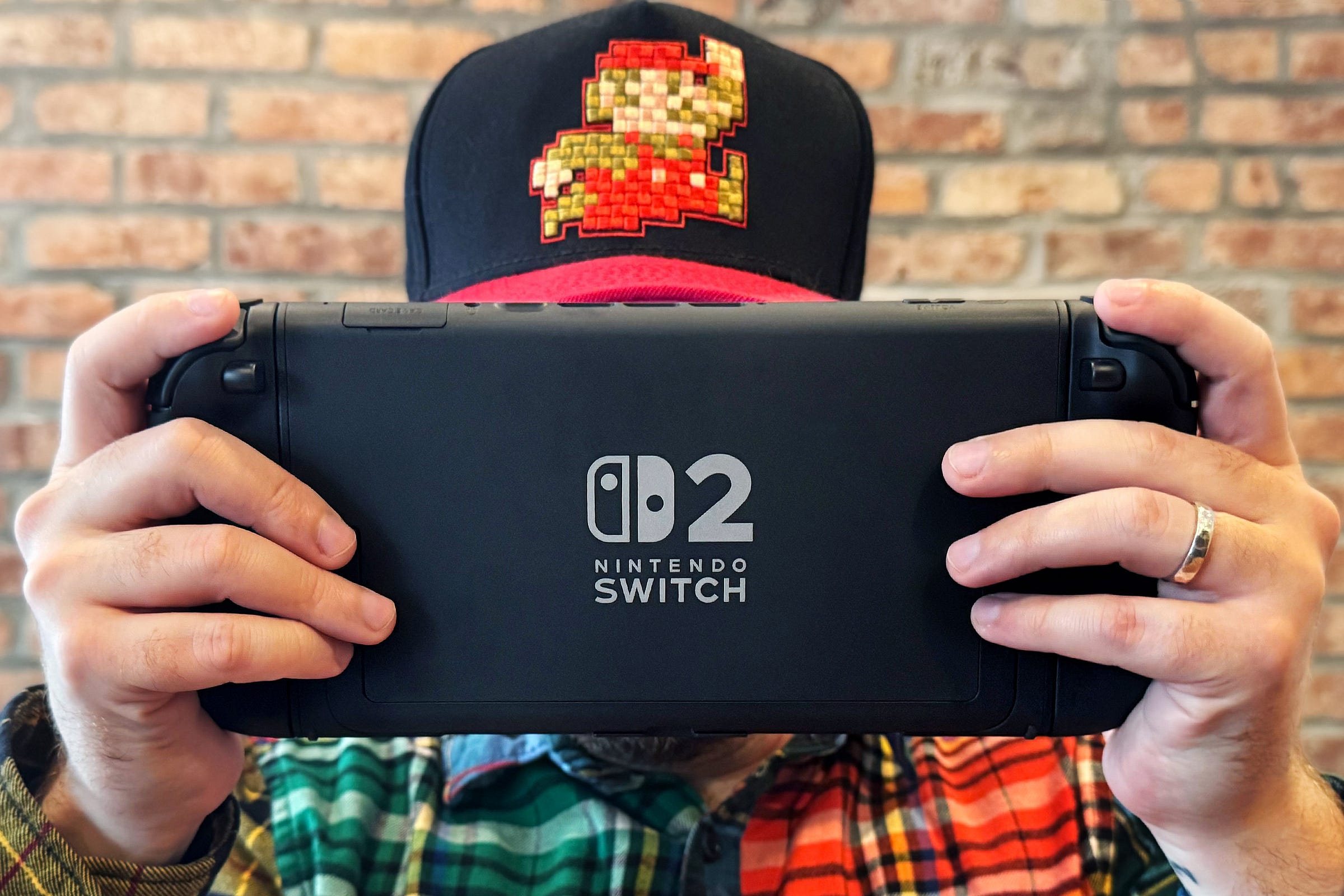 Color photograph of a man wearing a Mario baseball hat and holding the Nintendo Switch 2 in front of his face while he plays video games