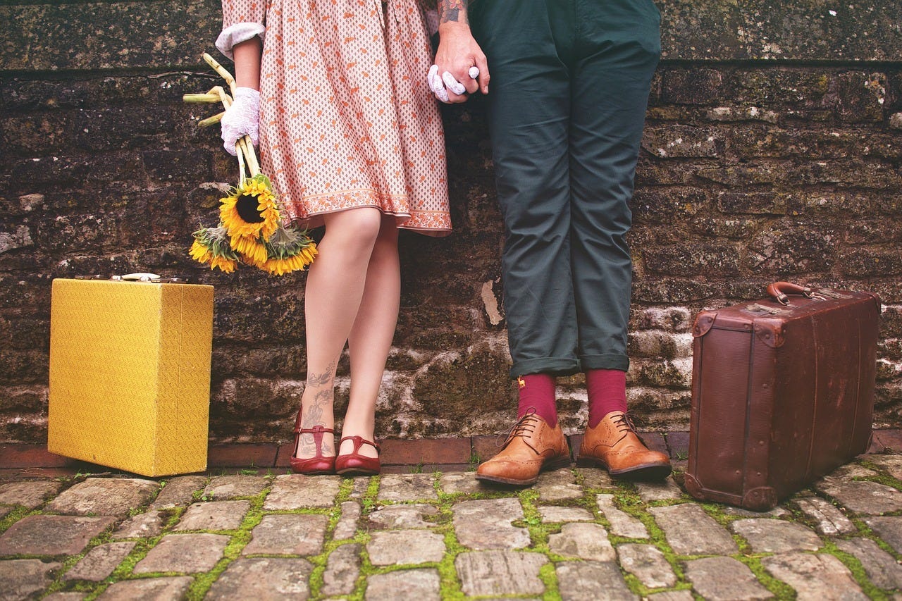 Light-skinned woman holding a bunch of sunflowers, wearing a red and white dress, white gloves, and red dressy shoes, standing next to a yellow vintage suitcase, holding hands with a light-skinned masculine-looking person wearing navy blue pants, red socks, and sable short boots, standing next to a brown vintage suitcase. They both have visible tattoos on ankle and wrist, and are standing against a brown brick wall, on white cobblestones with moss in the cracks. Light-skinned woman holding a bunch of sunflowers, wearing a red and white dress, white gloves, and red dressy shoes, standing next to a yellow vintage suitcase, holding hands with a light-skinned masculine-looking person wearing navy blue pants, red socks, and sable short boots, standing next to a brown vintage suitcase. They both have visible tattoos on ankle and wrist, and are standing against a brown brick wall, on white cobblestones with moss in the cracks.
