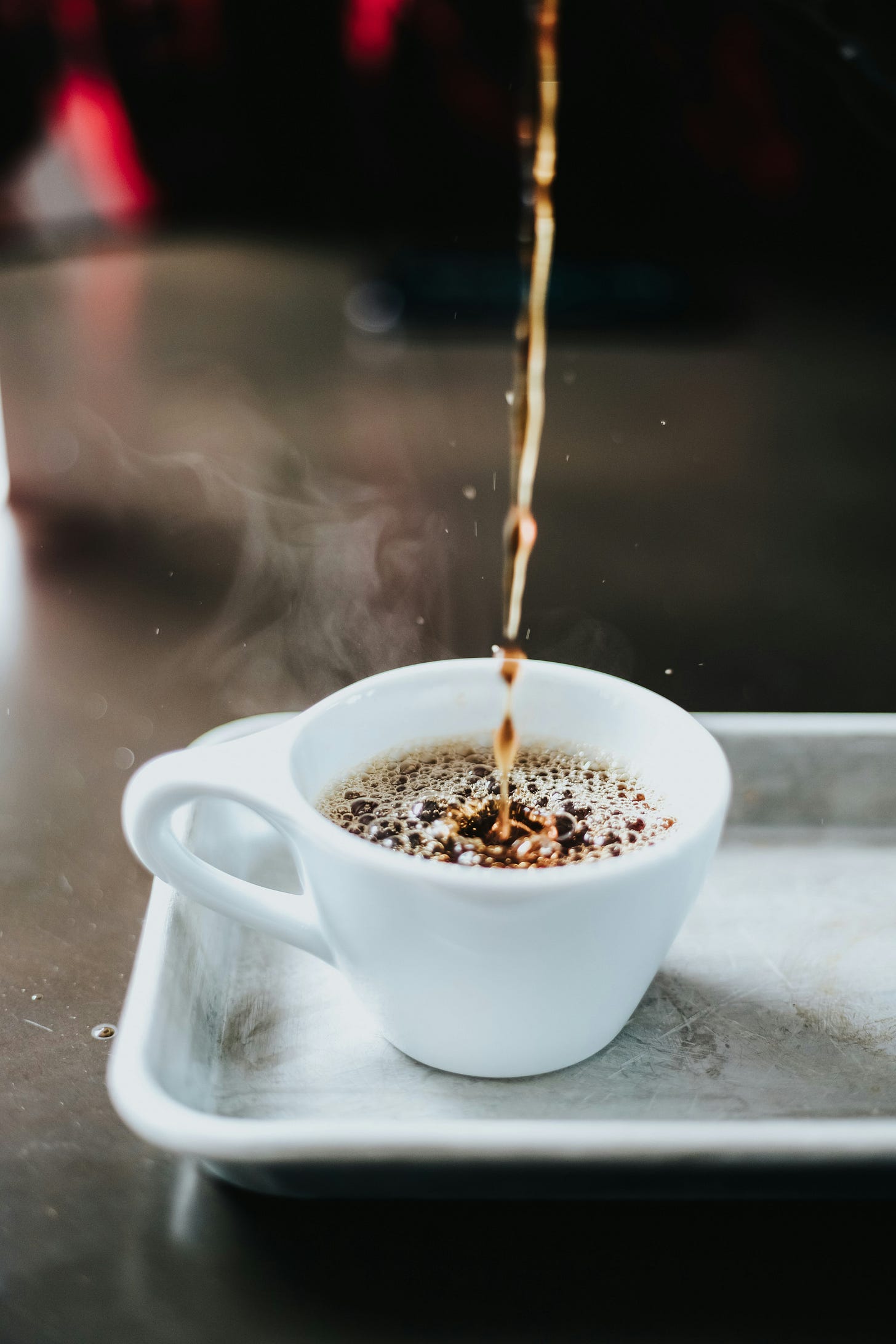 A stream of hot coffee is pouring into a white cup from a height. The cup sits on a silver tray, which rests on a table.
