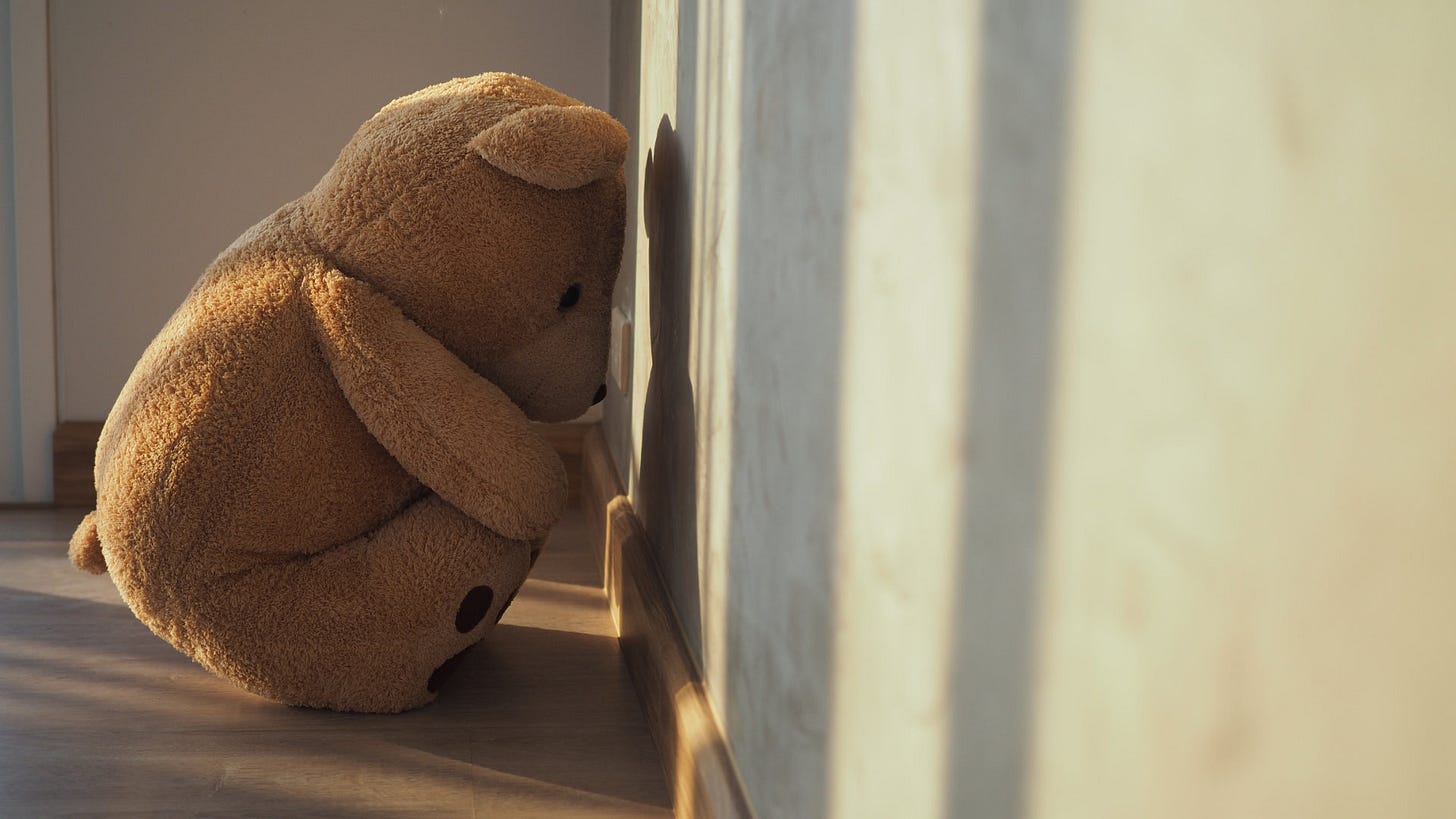 A teddy bear looking sad while leaning into a wall.