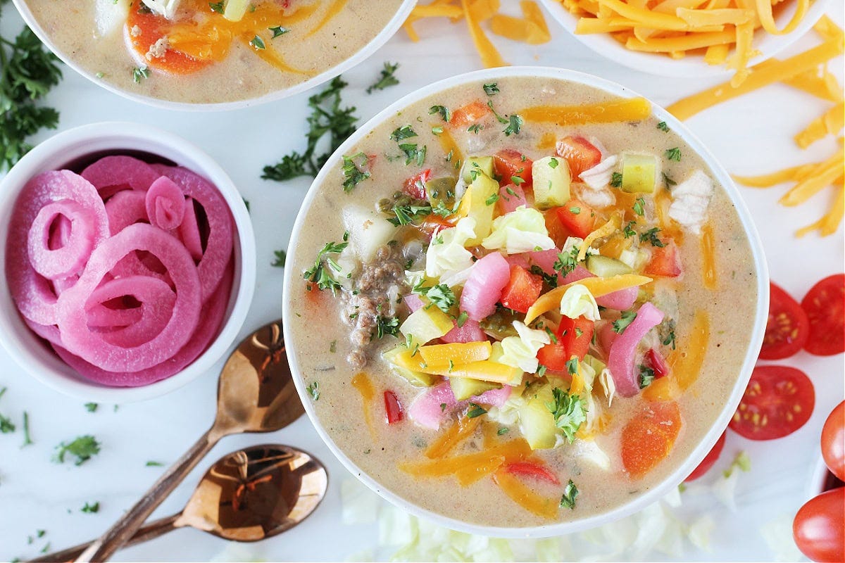 A bowl of cheesy soup topped with pickled onions, pickles and cheese.