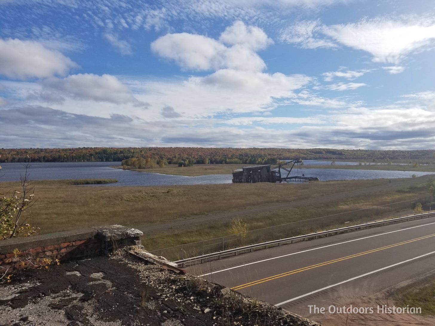 The photo is taken from an old building and looks across a road at an old dredge on shore of an inland lake. 