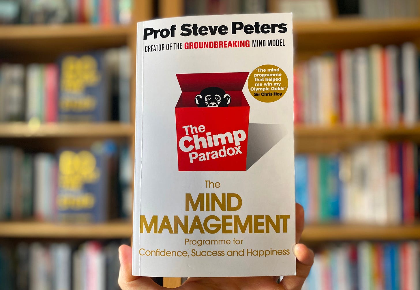 A photo of the book, the Chimp Paradox  by Prof Steve Peters in front of a set of bookshelves. The cover is white with a red box in the centre with a chimp's head poking out.