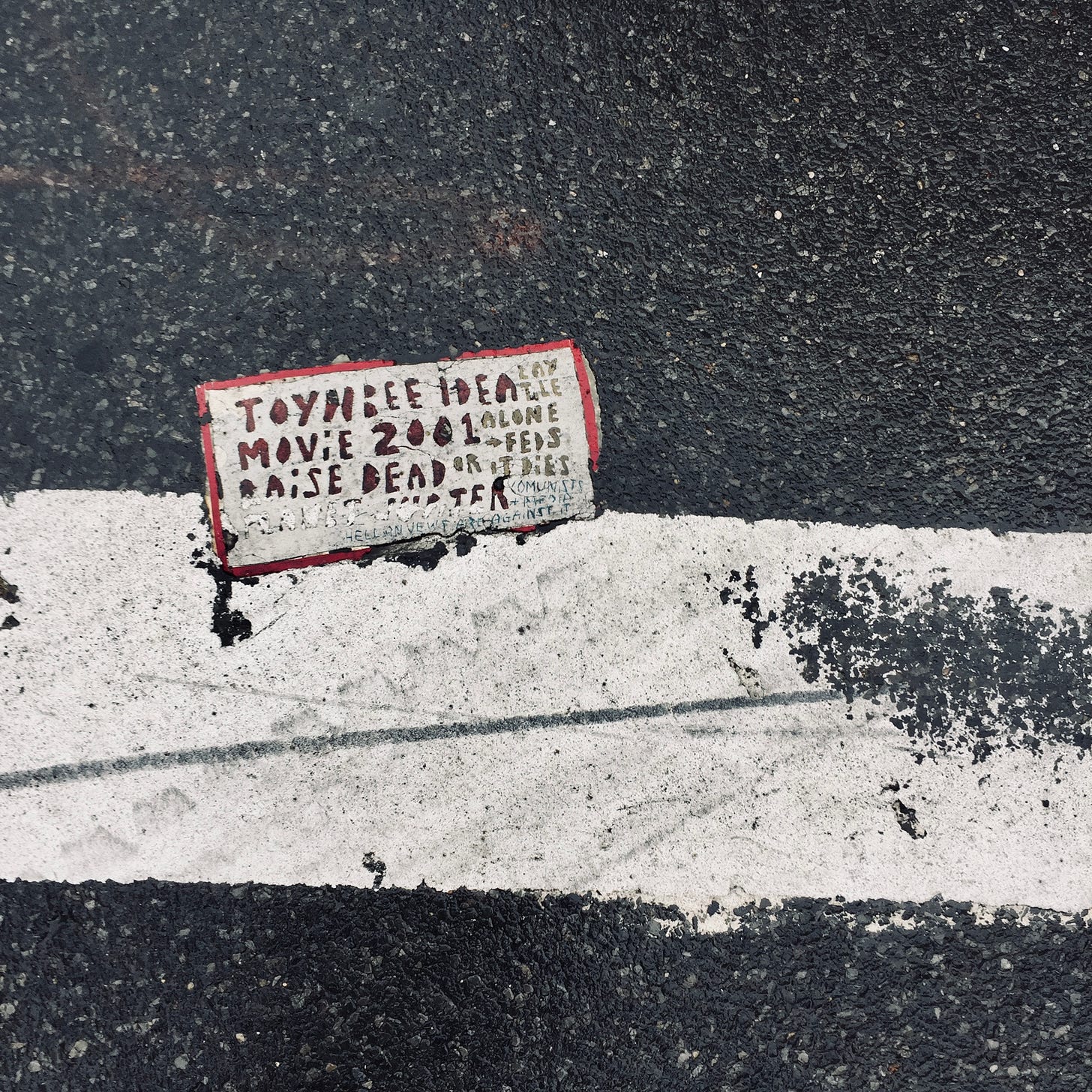 A mysterious tile in a crosswalk that partially reads TOYNBEE IDEA MOVIE 2001 RAISE DEAD
