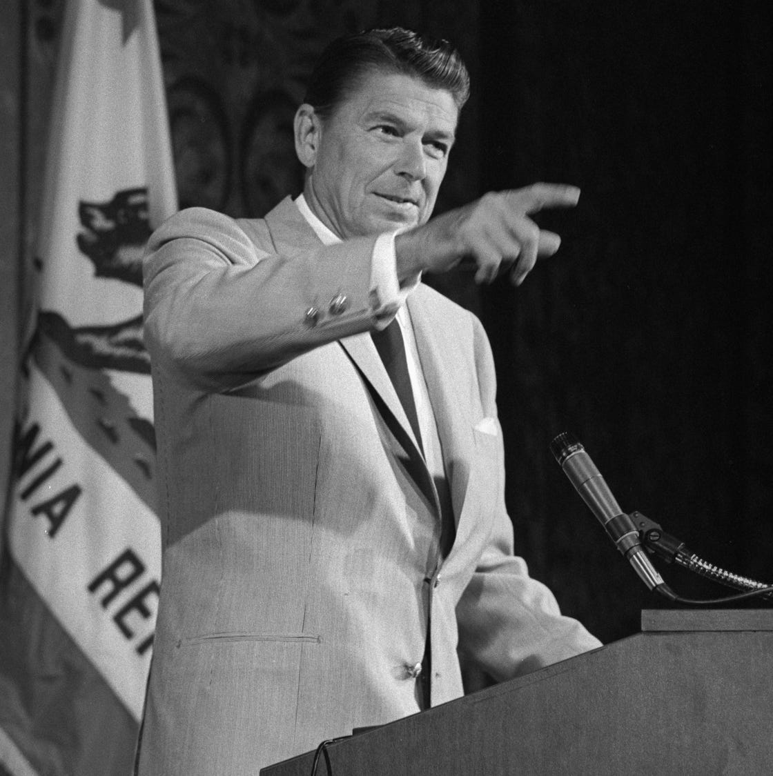 ronald reagan pointing as he stands at a podium with a california flag behind him