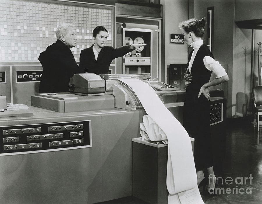 Black and white still from the film Desk Set (1957) in which IBM efficiency expert RIchard Sumner looks on as electronic brain operator Miss Warriner scolds librarian Bunny Watson in front of the room-sized computer, EMERAC.