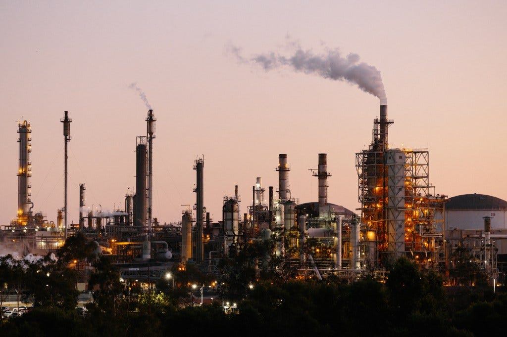 A wide view of a refinery location during sunset with various structures, including distillation columns, as smoke is released into the air.
